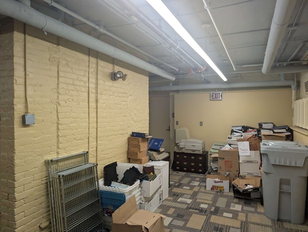 A small room with yellow walls. A pile of miscellaneous equipment lines its left side, and in the back is a pile of cardboard boxes full of three-ring binders, labelled TRASH.