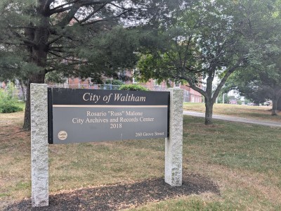 In front of a red brick building, a grove of trees and a sign identifying the property as the City of Waltham's Rosario "Russ" Malone City Archives and Records Center at 260 Grove Street.