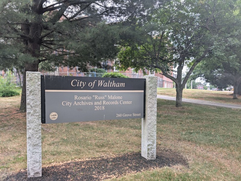 In front of a red brick building, a grove of trees and a sign identifying the property as the City of Waltham's Rosario "Russ" Malone City Archives and Records Center at 260 Grove Street.