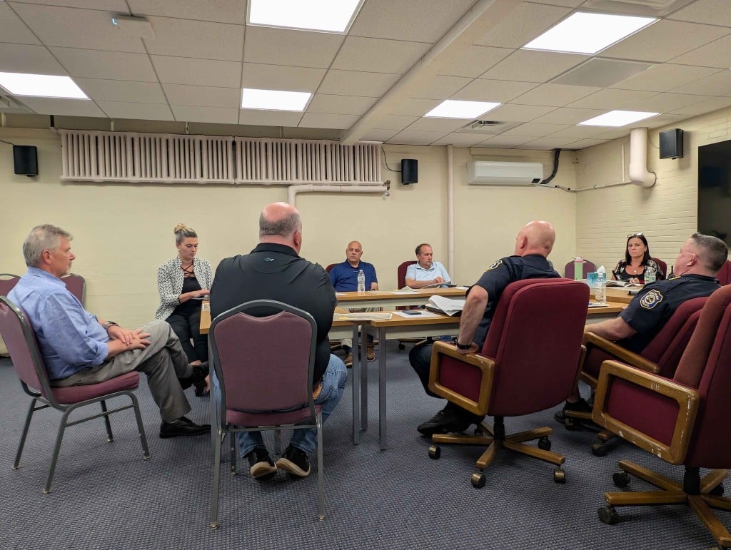 A group of Waltham officials and architectural planners sit around a conference table in the City Hall basement meeting room. The table includes City Councilors Paul S. Katz and Bill Hanley, Police Chief Kevin O'Connell, Deputy Police Chief Steven Champeon, Purchasing Agent Crystal Philpott, and principal project architect Jeffery McElravy.
